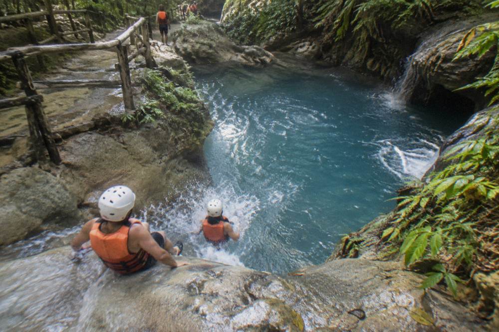 Small group in helmets and life vests sliding down a rock slide into a deep blue pool