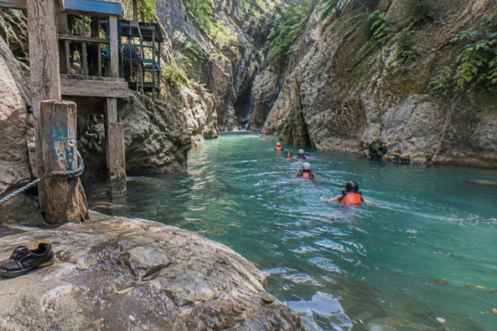 Group of visitors in helmets and life vests swimming across a wide turquoise pool between canyon walls